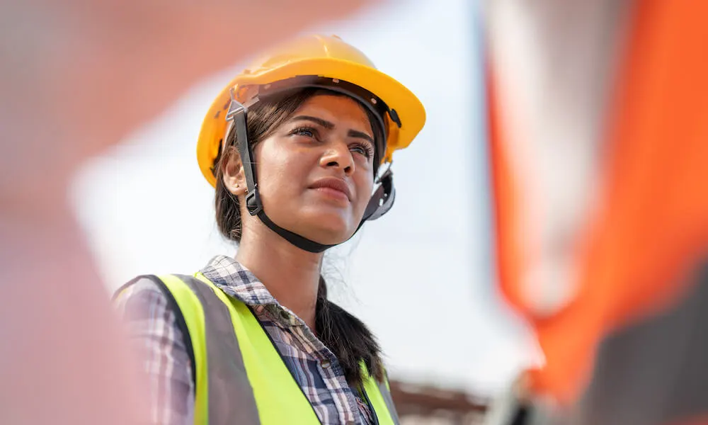An Indian Australian woman wears a hard hat and looks thoughtfully to something not in the photo 